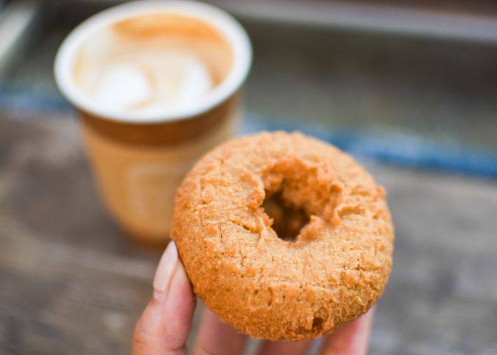 A small donut held up to the camera. In the background, a fresh cup of coffee to go.